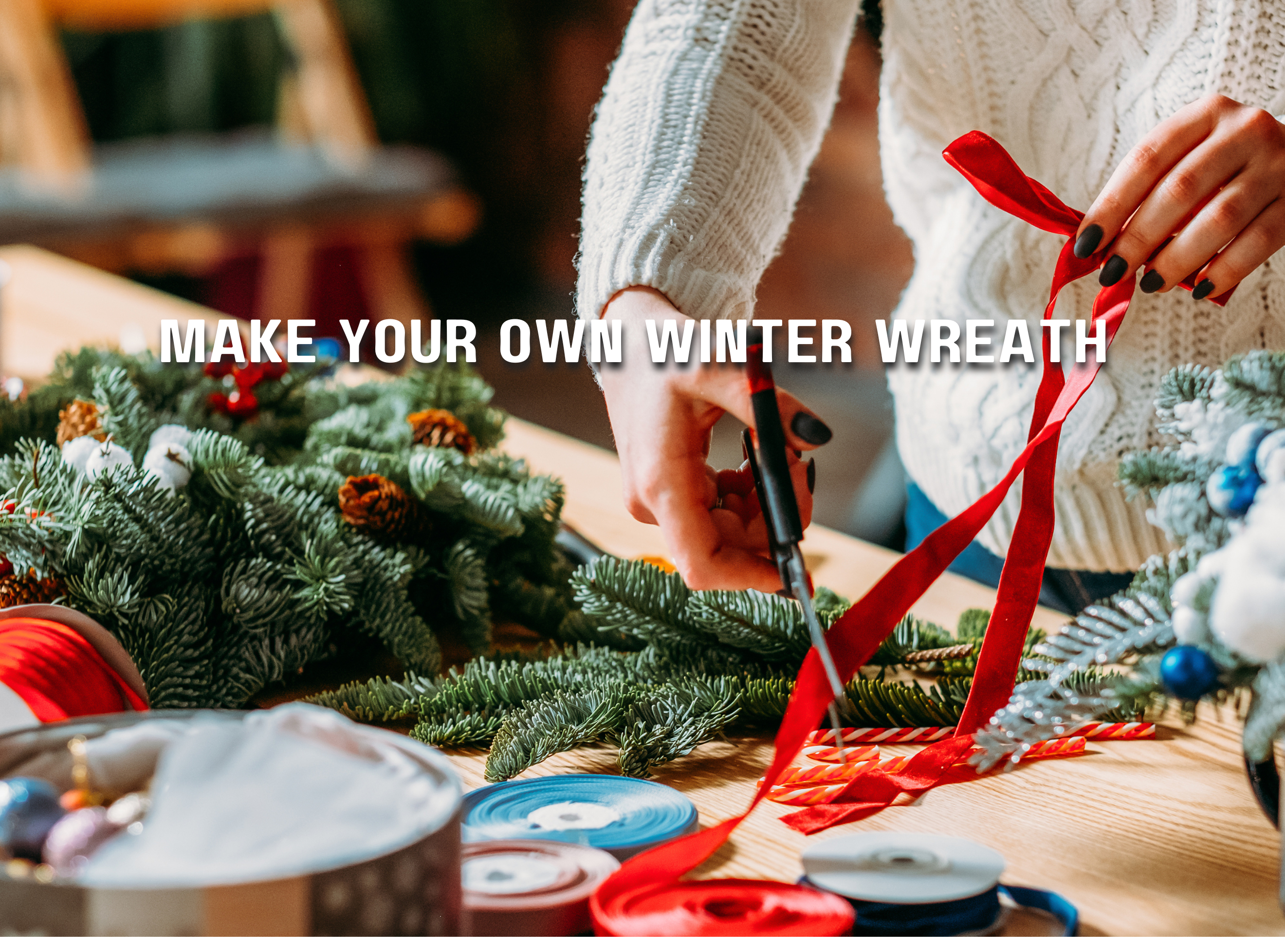 Participant is creating an evergreen winter wreath at a desk by cutting festive red ribbon.