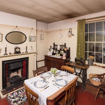 A Victorian Living room with a dining room table sitting at the centre with 4 places made. An oval mirror hangs above the fireplace.